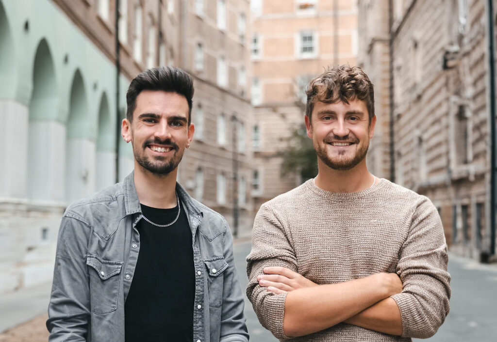 Two caucasian men stand smiling towards camera on a street in london