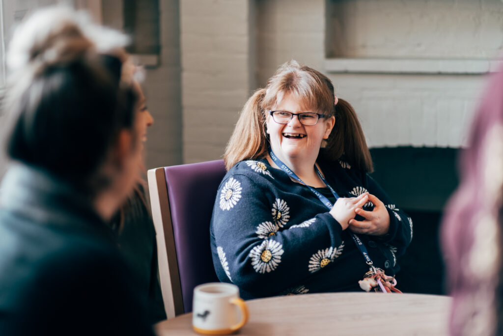 A resident at a coffee table smiling warmly at a staff member, captured from an over-the-shoulder perspective of the staff member.